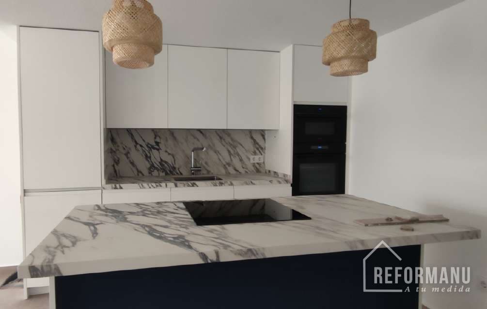 Cocina reformada por REFORMANU en Elche: isla y encimera de mármol, frente blanco e iluminación de diseño